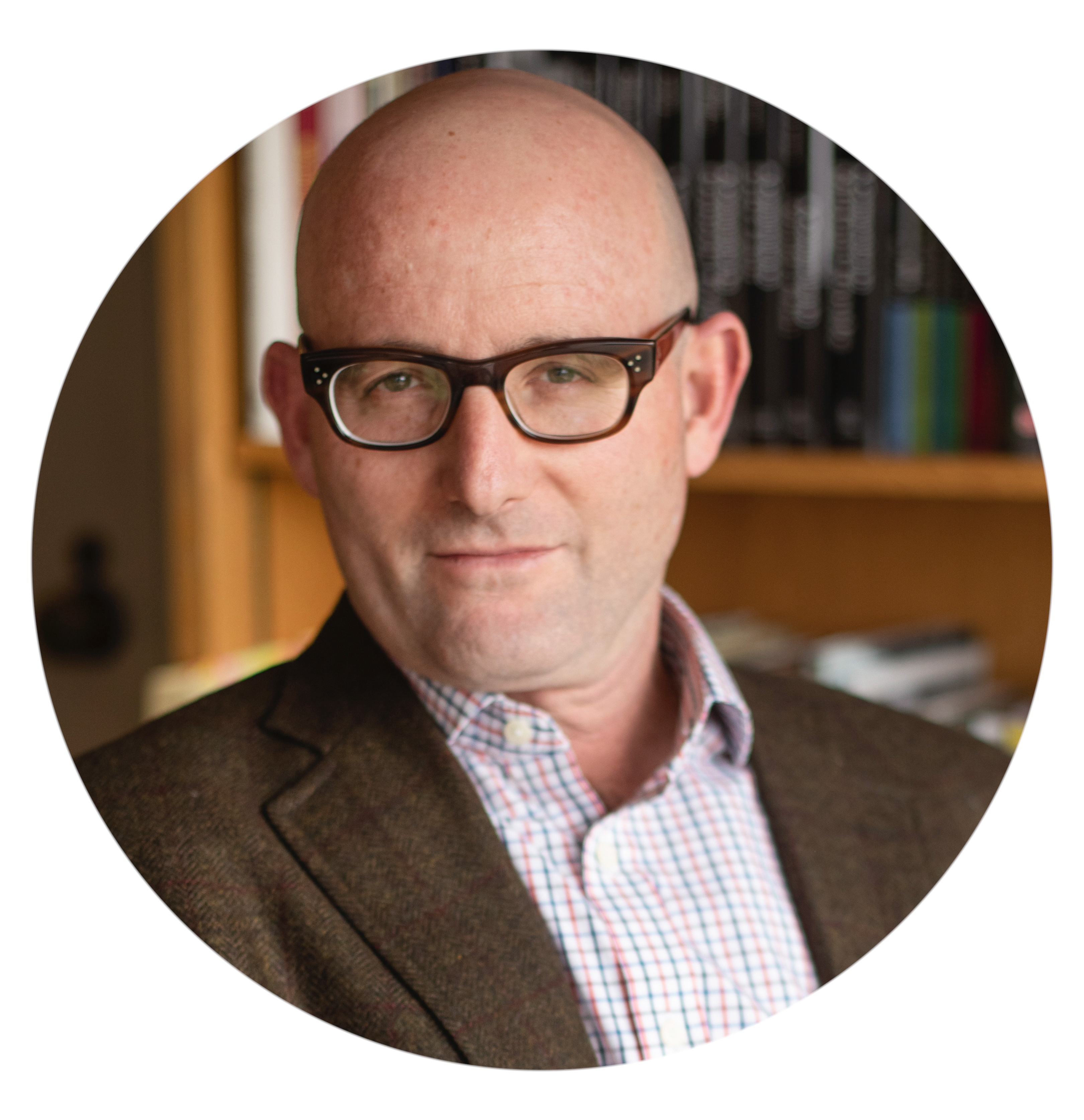 headshot of a bald man wearing black square glasses and a brown jacket over an open collared shirt